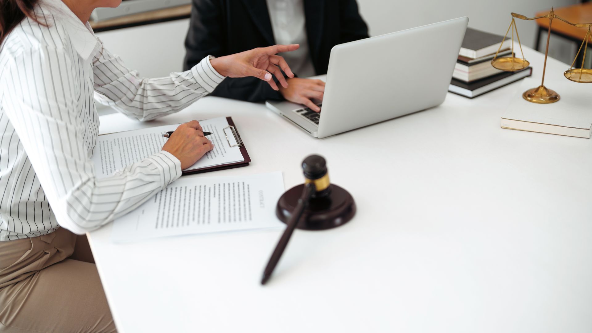 Une personne en costume examine un document avec une autre personne assise à une table, un ordinateur portable est présent.