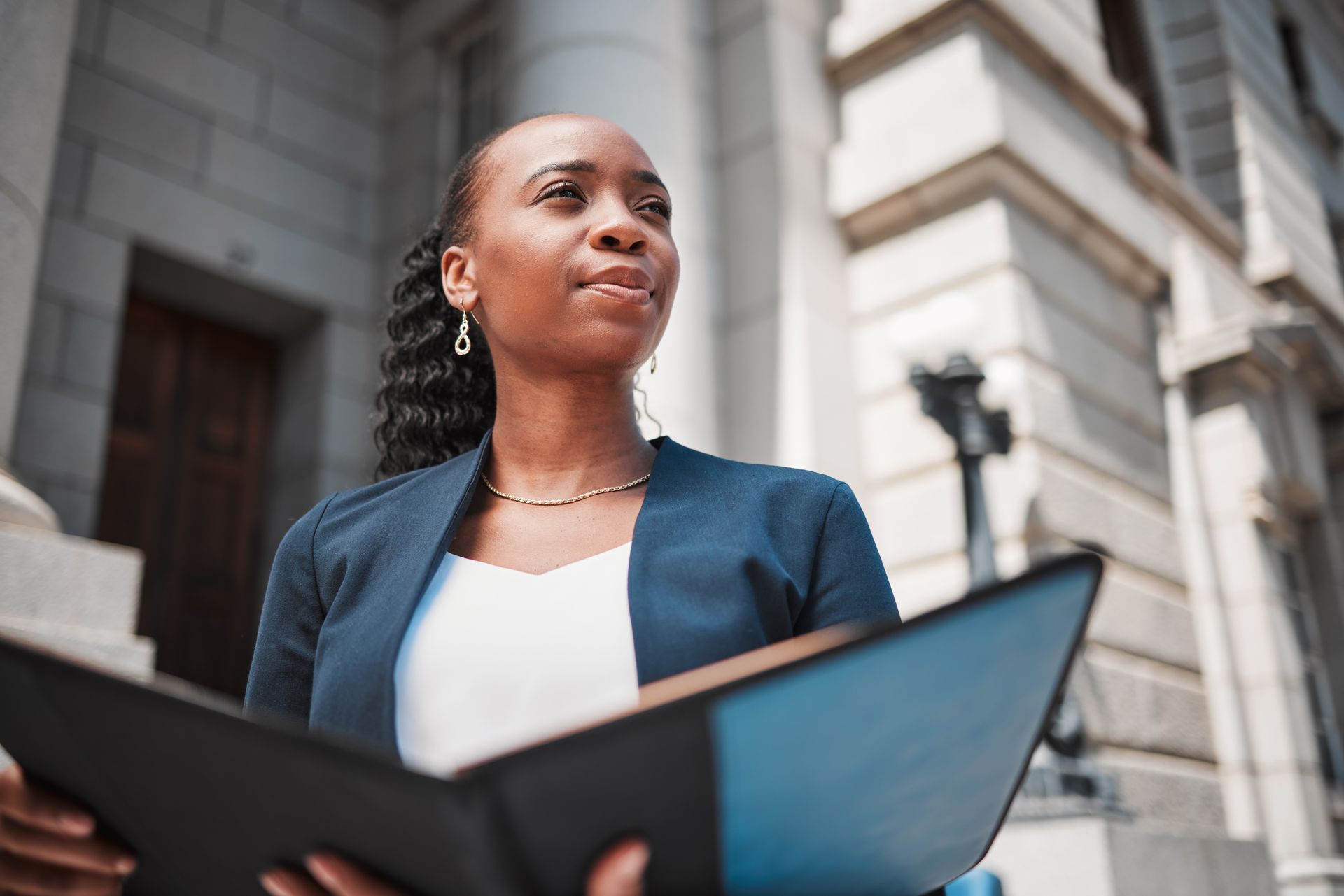 Une femme en blazer, tenant des documents, se tient devant un bâtiment, le regard levé vers le ciel avec une expression pleine d'espoir.