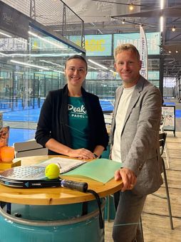 Een vrouw en een man staan lachend aan een tafel. Achter hen zijn de padelbanen zichtbaar.