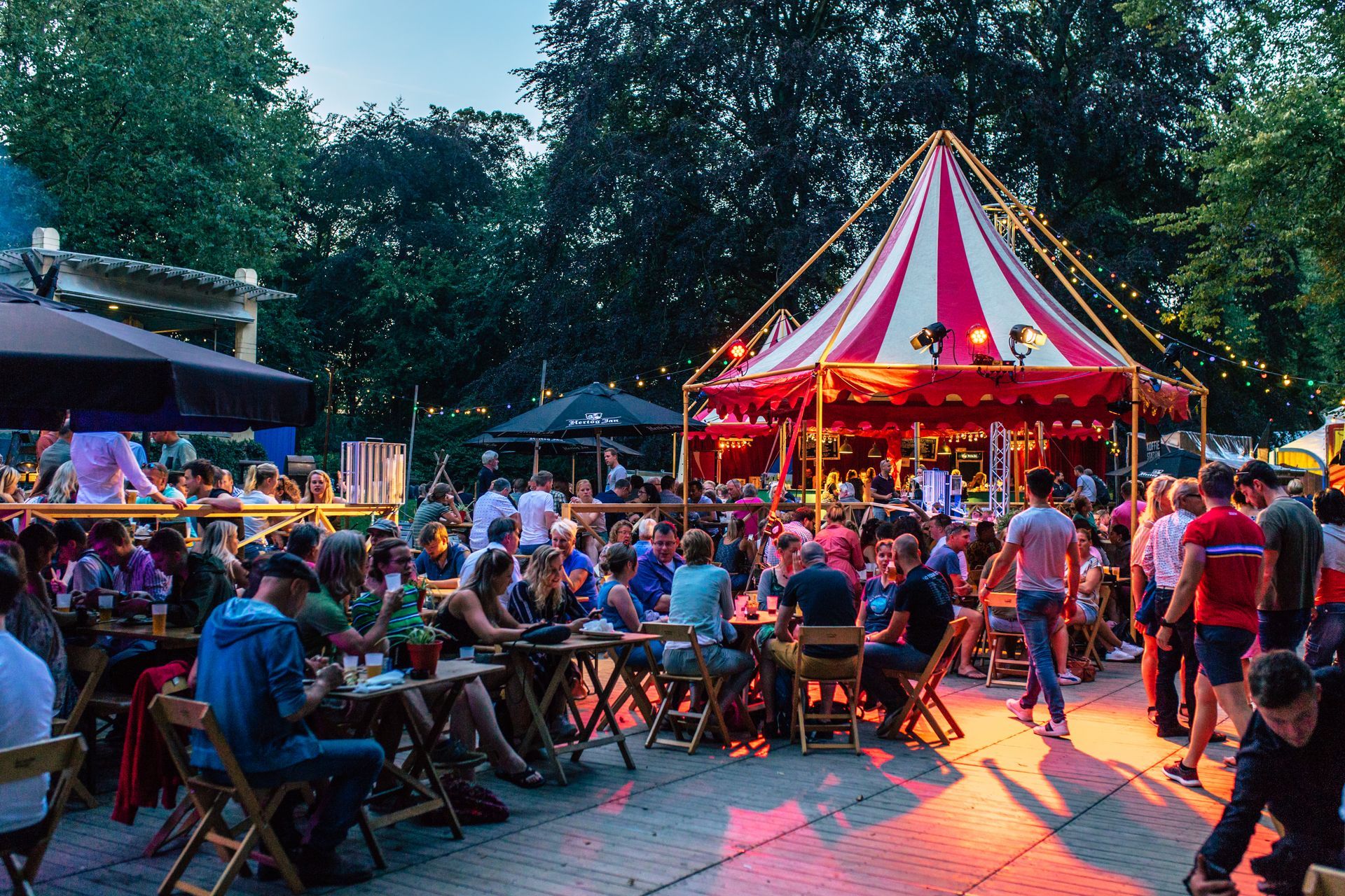 Avondbijeenkomst in de buitenlucht met mensen aan tafels, onder een circustent. 