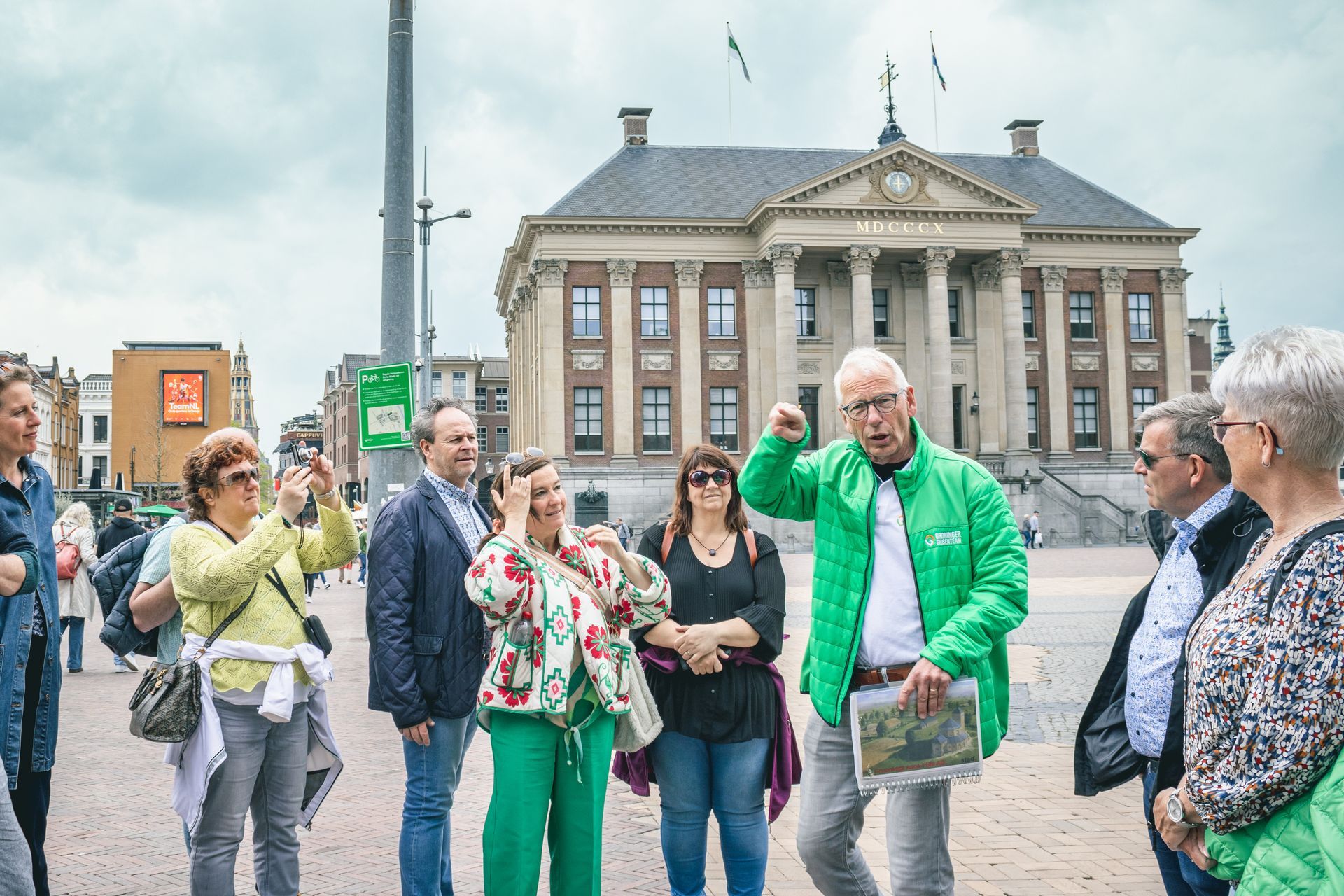 Een gids van het Groninger Gidsenteam wijst naar een gebouw. ​​Een groep mensen staat eromheen en maakt foto's.