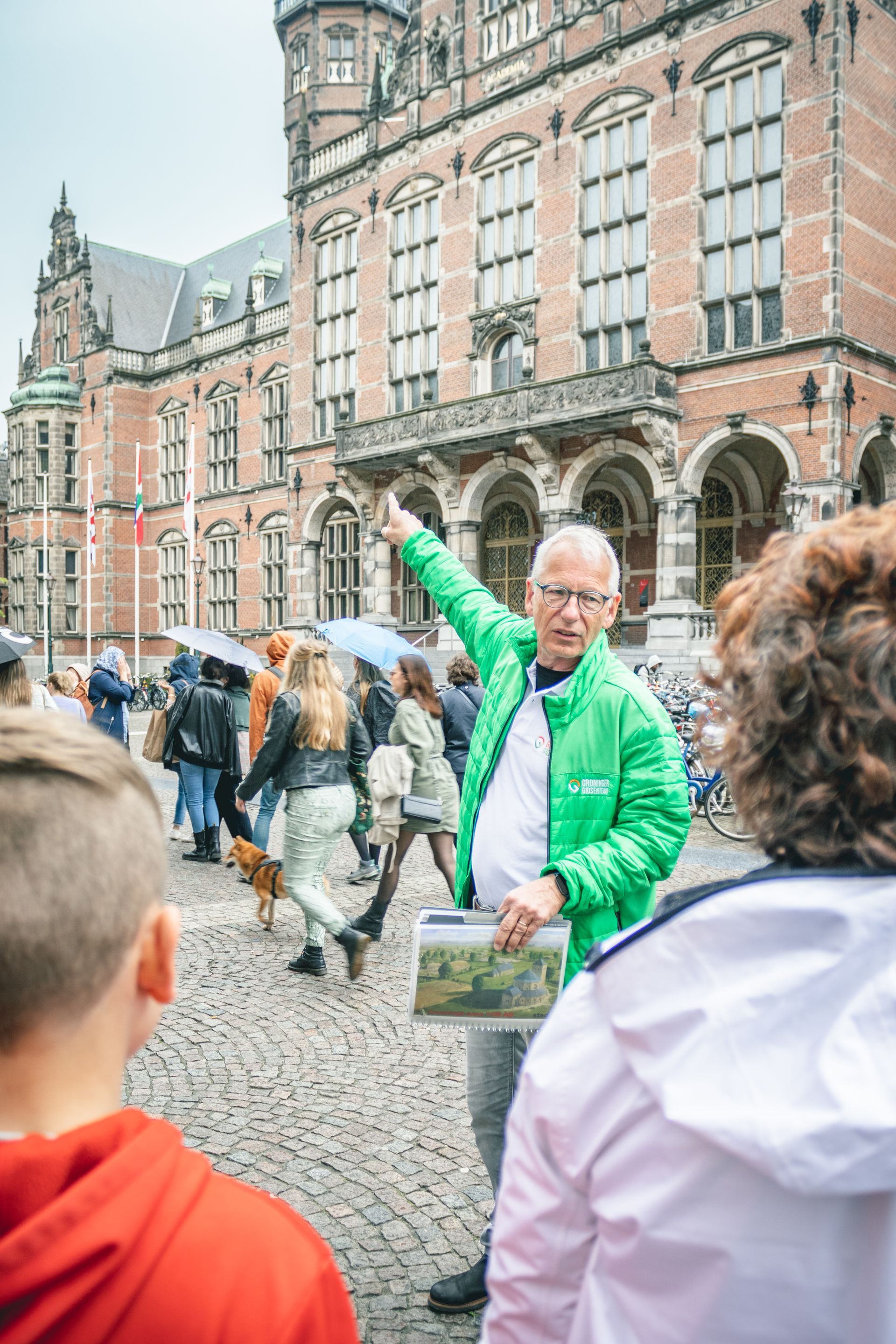 De gids van Groniger gidsenteam wijst naar station Groningen. ​​Een groep mensen luistert. Het is een lichtbewolkte dag 