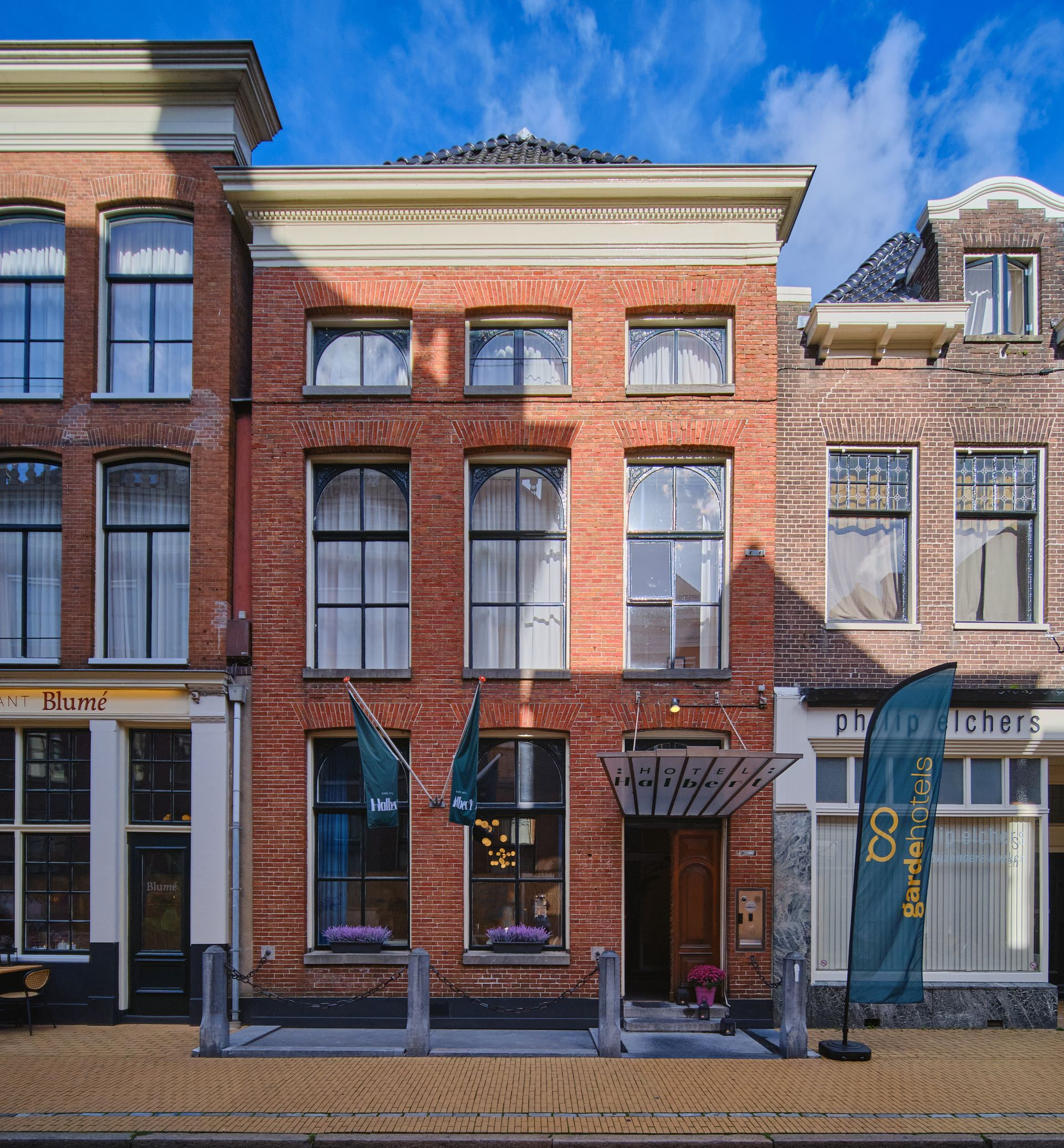 De bakstenen gevel met hoge ramen in de Oude Boteringestraat van Hotel Halbert. Een deuropening onder een luifel, vlaggen en uithangborden van Gardehotels aan een zonnige straat.