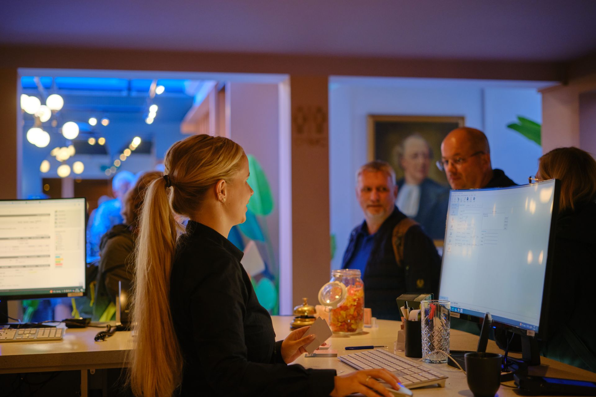 Een man achter een computer staat lachend aan de balie met een laptop en iemand anders achter hem.