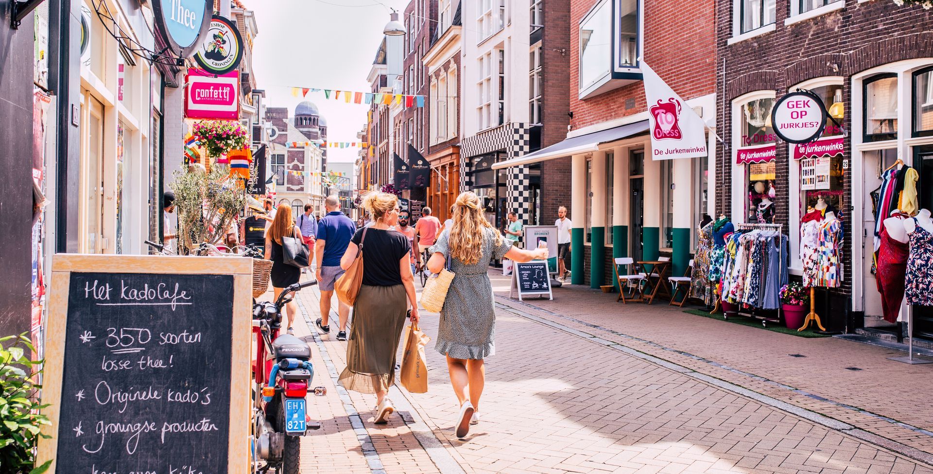 Straat met winkels en wandelende mensen. Twee vrouwen in jurken slenteren door de straat op een zonnige dag.