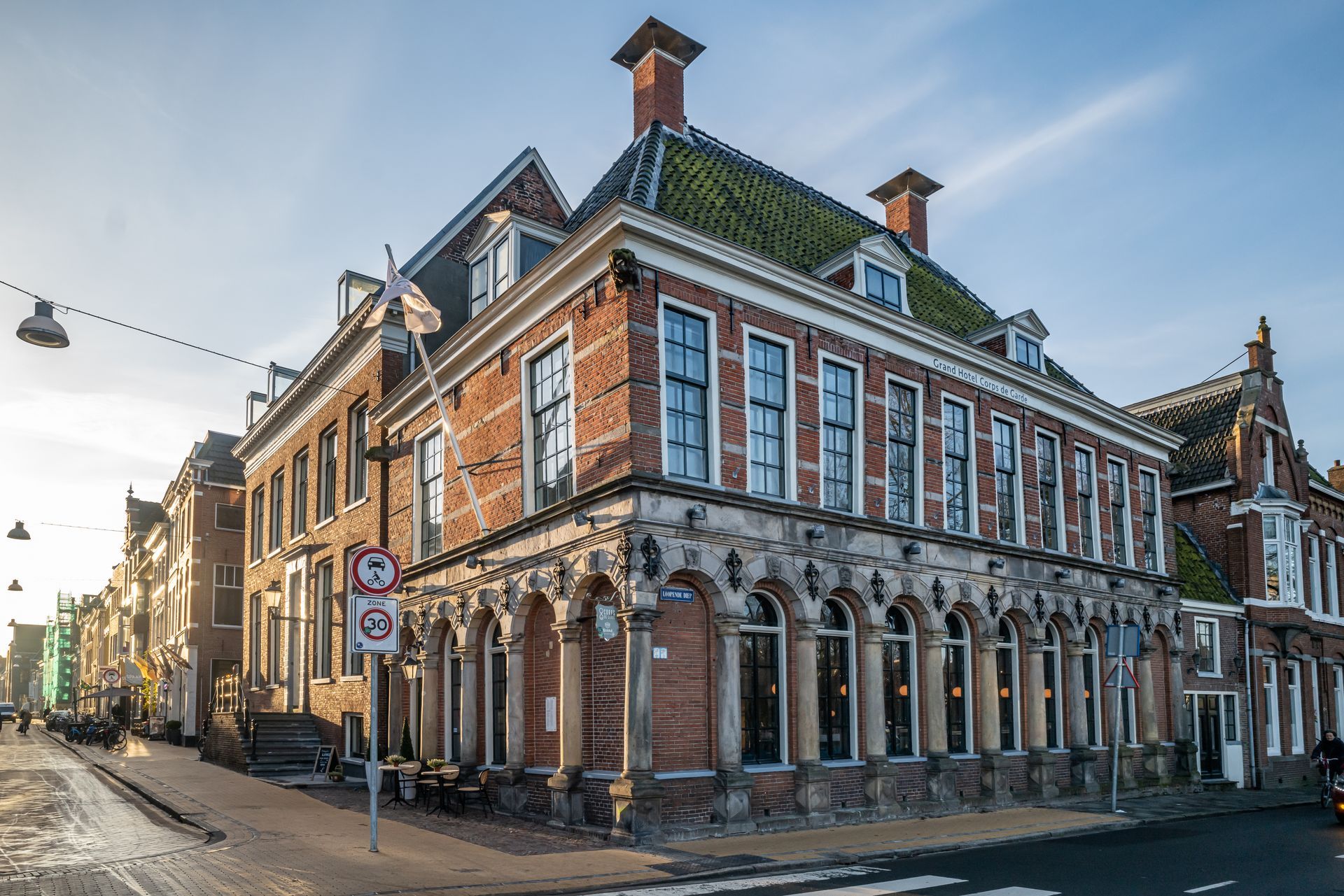 Gebouw van rode baksteen met boogramen, vlag en uitzicht op straat onder een bewolkte hemel.