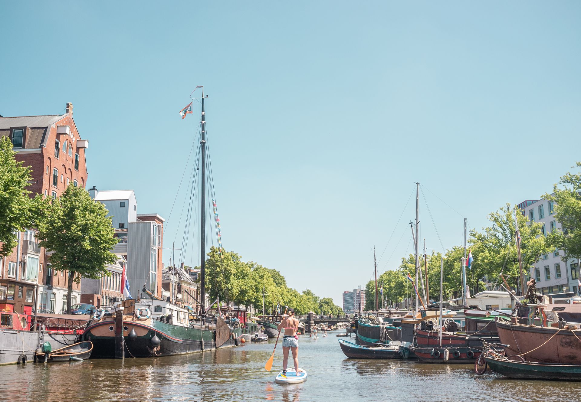 Een persoon op een paddleboard in een kanaal, omringd door boten en gebouwen onder een stralendblauwe lucht.