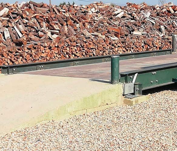 Pila de llenya sobre una plataforma de formigó, amb grava en primer pla i suports metàl·lics.