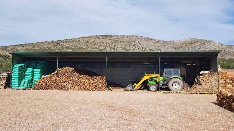 Un tractor amb carregador frontal es troba sota un cobert ple de llenya i palets, davant d'una muntanya.