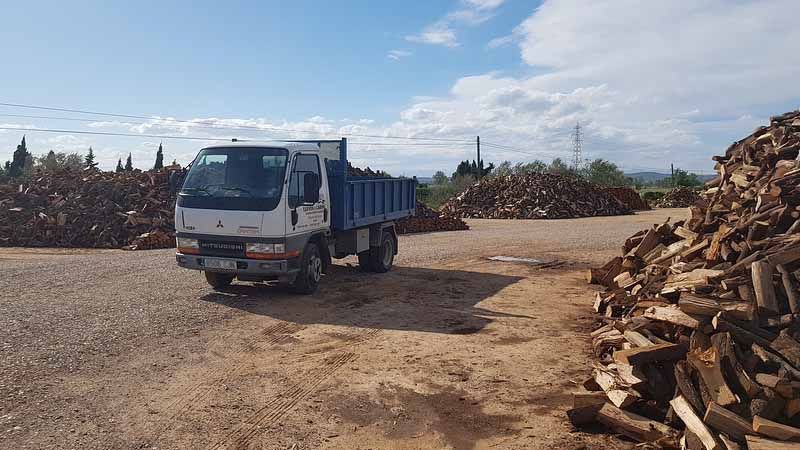 Camió carregat de llenya, aparcat en un magatzem de fusta sota un cel blau, amb piles de troncs a banda i banda.