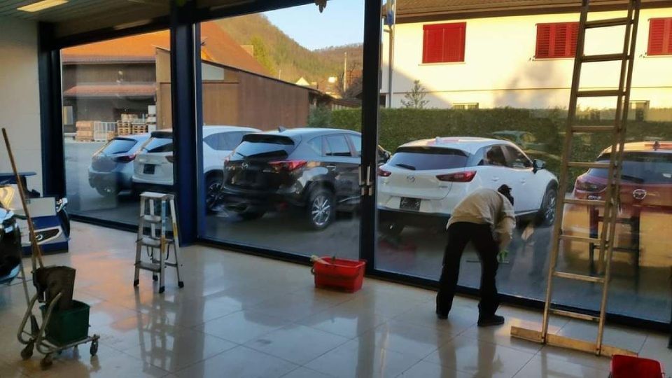 Eine Person reinigt ein grosses Schaufenster in einem Autohaus. Durch das Fenster sind Autos zu sehen.