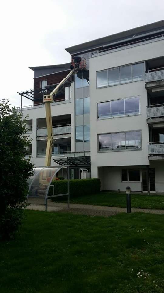 Eine Person arbeitet in einem Aufzug auf dem Dach eines weissen Wohnhauses. Grüner Rasen und bedeckter Himmel.