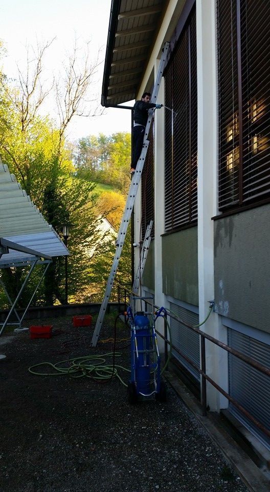 Eine Person reinigt auf einer Leiter die Aussenfassade eines Gebäudes. Das Gebäude hat braune Fensterläden und steht neben einem Baum.