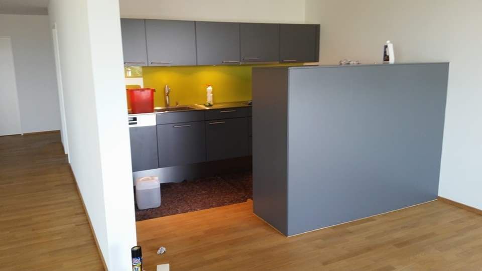 Graue Küche mit Ober- und Unterschränken, braunem Bodenbelag und einer Trennwand.