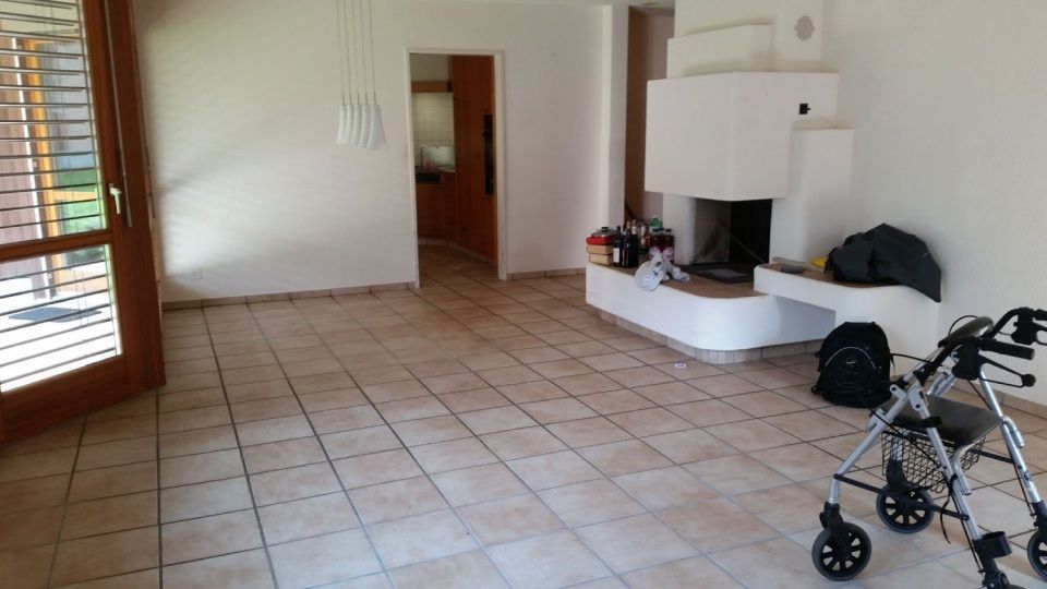 Leeres Zimmer mit Fliesenboden, weissem Kamin, offener Tür und Rollator.