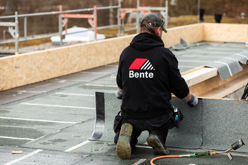 Eine Person verlegt Dachmaterial mit einem Brenner. Das gerollte Material ist schwarz, und Flammen sind sichtbar.