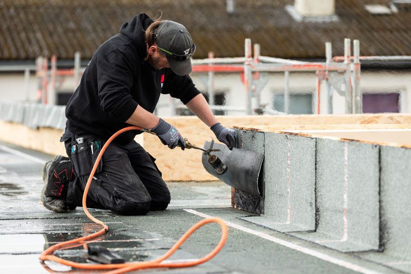 Mann auf einer Leiter arbeitet an einer Dachrinne. Er hält Werkzeug und ein Rohr. Im Hintergrund eine Backsteinmauer.