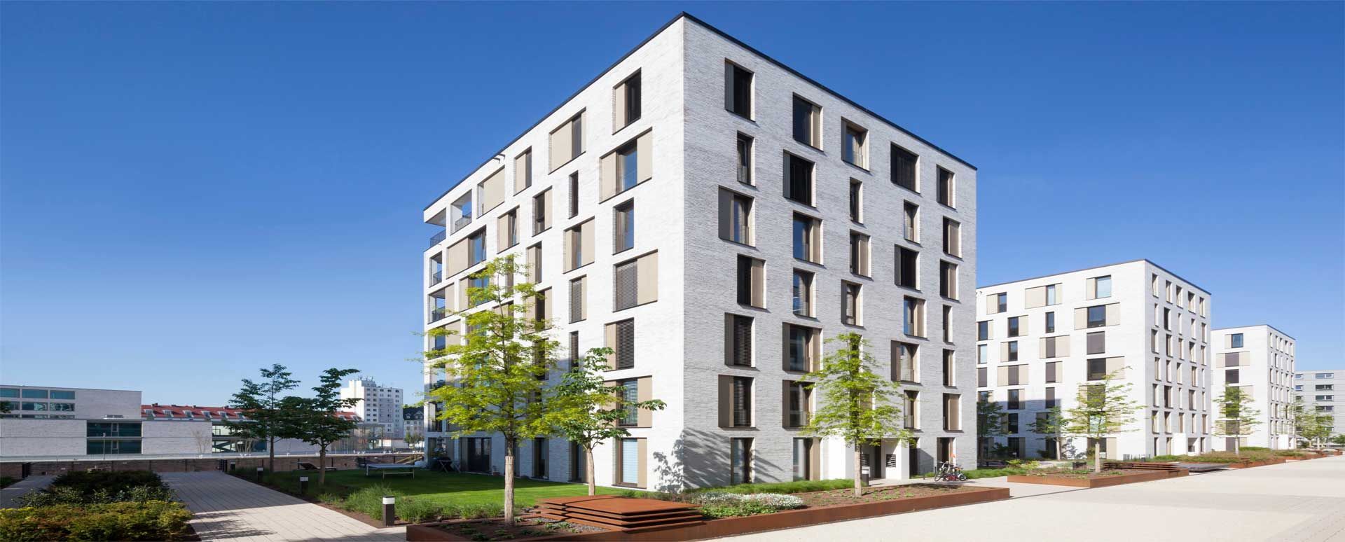 Moderne weiße Wohnhäuser unter klarem blauem Himmel, im Vordergrund Bäume und eine grüne Rasenfläche.