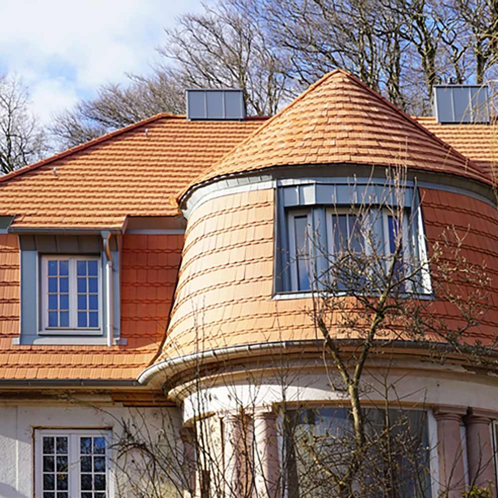 Das rotziegelgedeckte Dach eines Hauses mit abgerundetem Turm und rechteckigen Fenstern hebt sich vom blauen Himmel ab.
