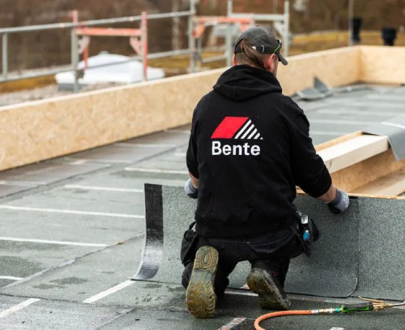 Mitarbeiter von Dachdecker Bente kniet auf einem Flachdach und arbeitet an der Dachbahn, im Hintergrund ein Baugerüst.