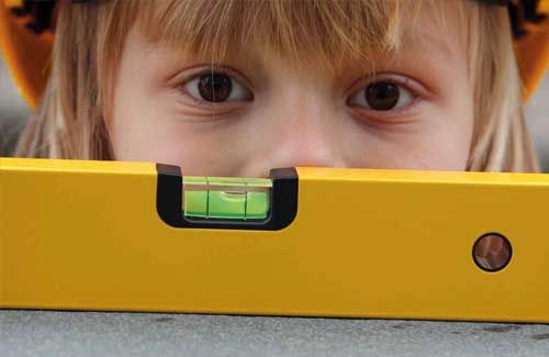 Kind schaut durch eine gelbe Wasserwaage. Trägt einen gelben Bauhelm. Nahaufnahme der Augen und der Wasserwaage.