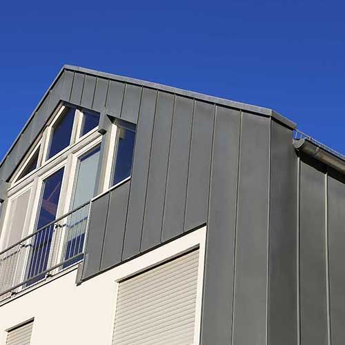 Graue Metallverkleidung an einem Gebäude mit Balkon vor strahlend blauem Himmel.