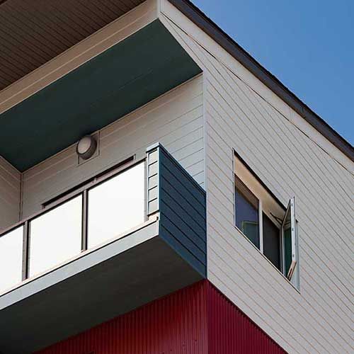 Modernes Eckgebäude mit Balkon und offenem Fenster. Weiße Fassade, türkisfarbene Zierelemente und rote Akzente vor blauem Himmel.