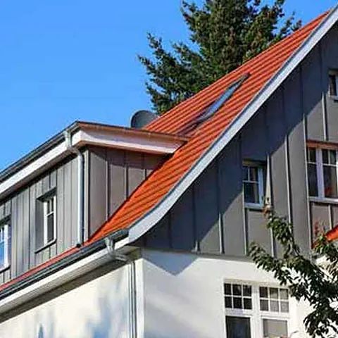 Haus mit rotem Dach, grauer Fassade, weißen Wänden und blauem Himmel im Hintergrund.