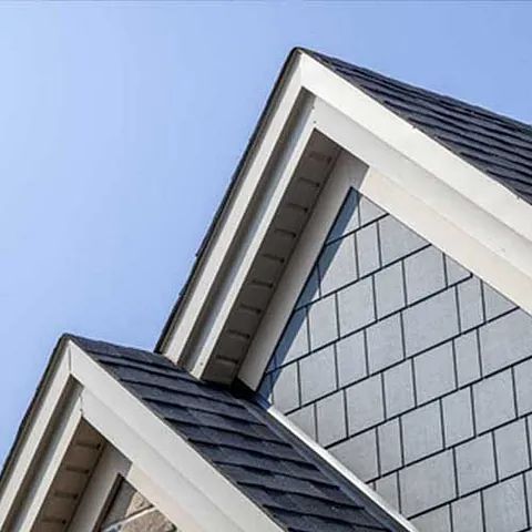 Nahaufnahme einer Hausdachecke mit dunklen Schindeln, weißen Zierleisten und hellblauer Fassadenverkleidung vor klarem Himmel.