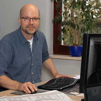 Mann mit Brille an einem Schreibtisch, der einen Computer benutzt. Blau kariertes Hemd. Fenster und Pflanzen im Hintergrund.