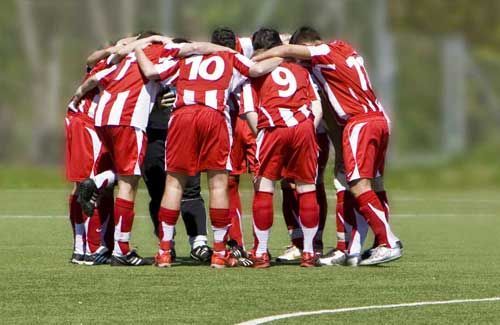 Die Fußballmannschaft versammelt sich auf einem grünen Feld. Die Spieler in rot-weiß gestreiften Trikots haben die Arme umeinander geschlungen.