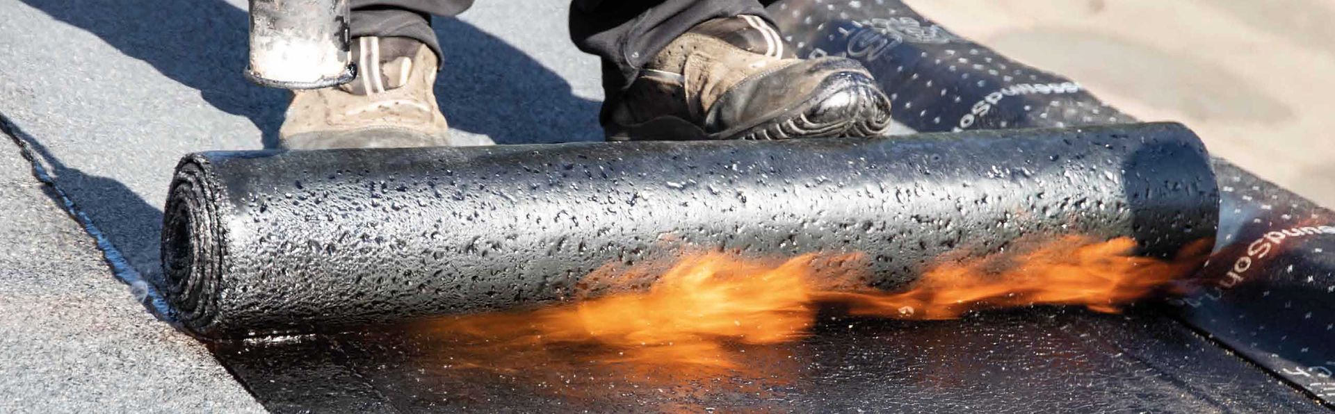 Eine Person verlegt Dachmaterial mit einem Brenner. Das gerollte Material ist schwarz, und Flammen sind sichtbar.