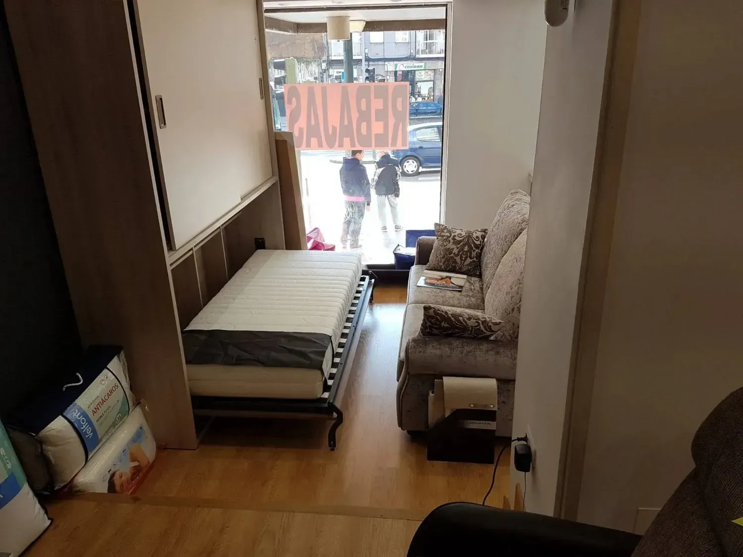 Una exposición que muestra una cama plegable abierta junto a un sillón estampado, con un escaparate al fondo.