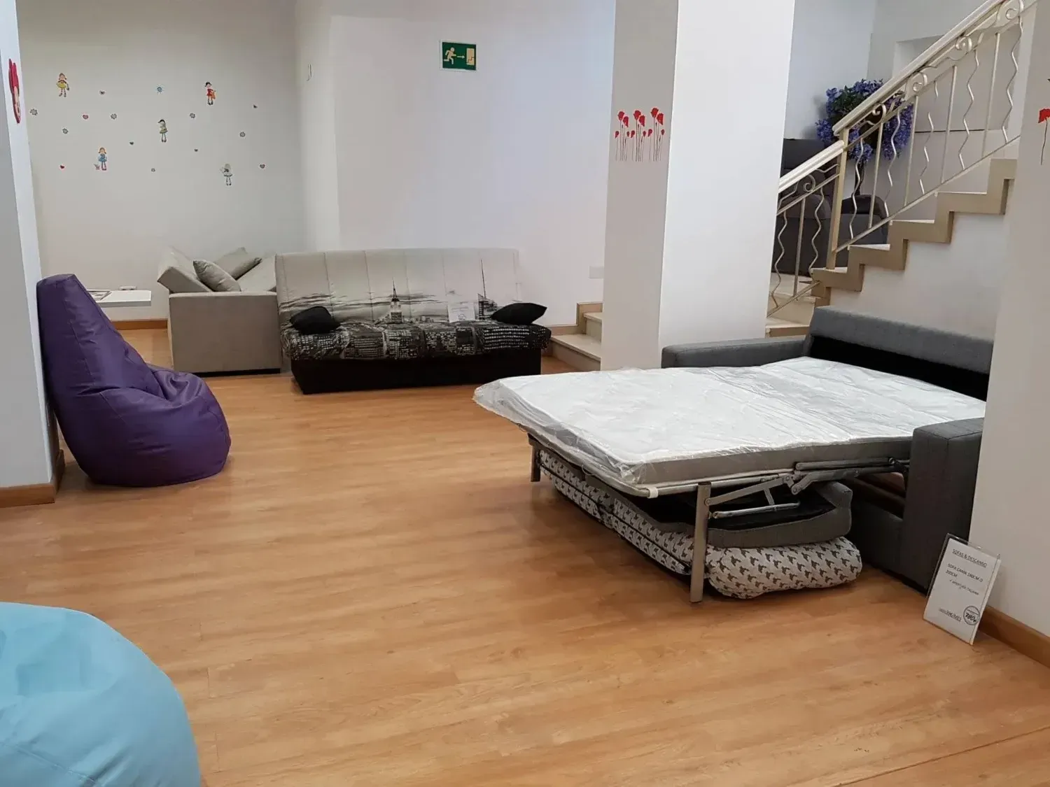 Una sala de exposición con varios sofás, incluyendo un sofá cama gris en primer plano, y coloridos pufs.