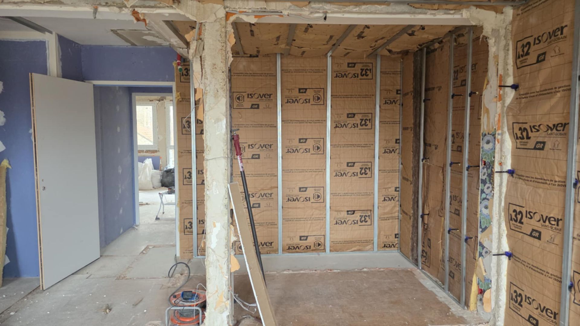 Aménagement intérieur : mur partiellement fini avec fenêtre, isolation, montants métalliques et escabeau.