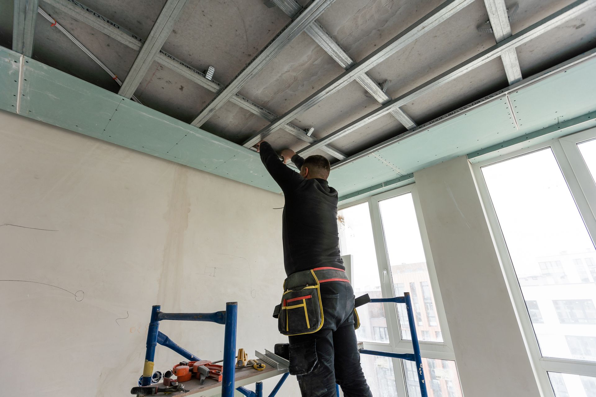 Un homme sur un échafaudage installe un plafond en plaques de plâtre dans une pièce lumineuse.