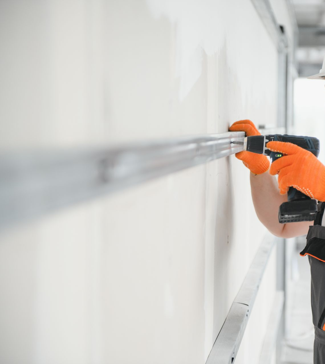 Une personne portant des gants orange installe un cadre métallique sur un mur en plaques de plâtre à l'aide d'une perceuse électrique.