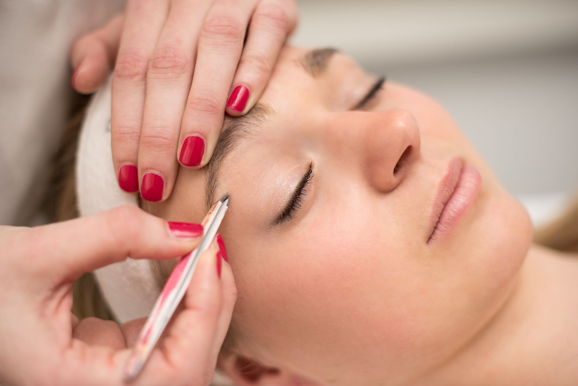 Épilation des sourcils avec une pince tenue par une main avec des ongles vernis de rouge