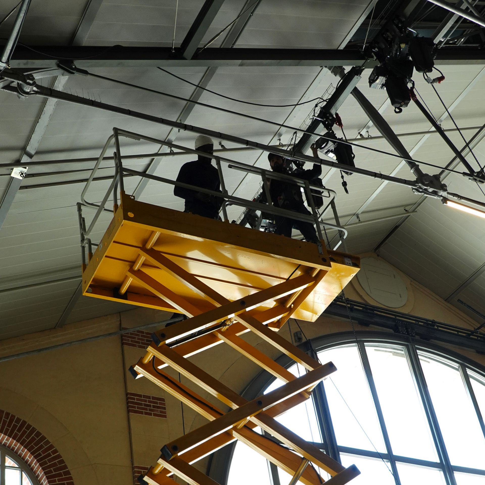 Des personnes se tiennent sur une nacelle élévatrice jaune à l'intérieur d'un grand bâtiment industriel, vue d'en bas.