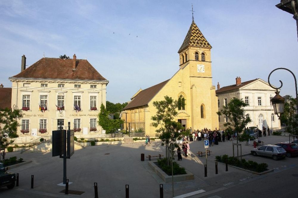 Place de village ensoleillée avec une église jaune, des bâtiments historiques et des gens rassemblés près des voitures garées
