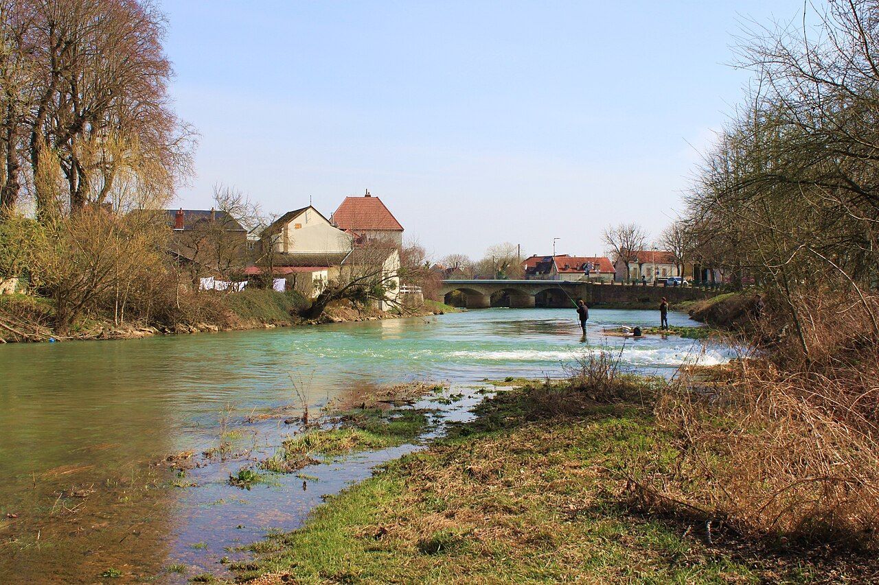 Paysage fluvial paisible avec des eaux vertes, des arbres dénudés, un petit pont et des maisons sous un ciel clair