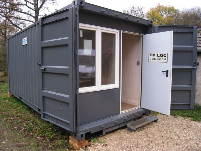 Conteneur de bureau portable gris foncé avec porte et fenêtres blanches ouvertes sur un terrain en gravier