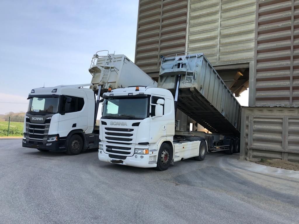 Deux semi-remorques blanches déchargent du grain dans un grand silo industriel.