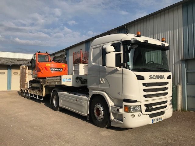 Camion-plateau Scania blanc transportant une excavatrice orange à côté d'un bâtiment industriel