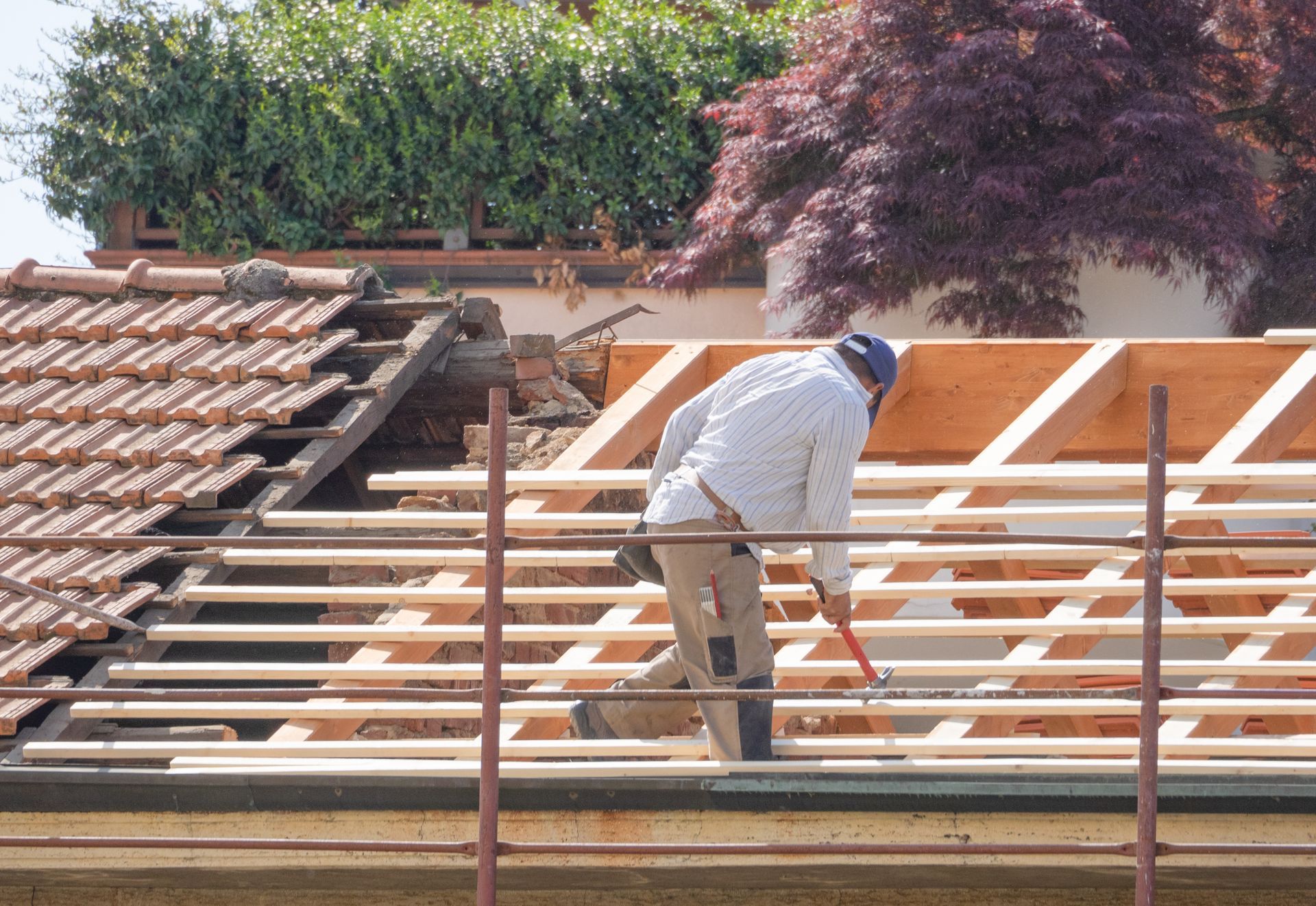 Un ouvrier installe des poutres de toiture sur une maison en construction, avec un toit de tuiles et des arbres en arrière-plan.