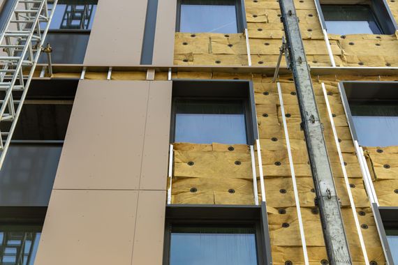 Façade d'un bâtiment en construction avec panneaux beiges, isolation apparente et cadres de fenêtres bleus