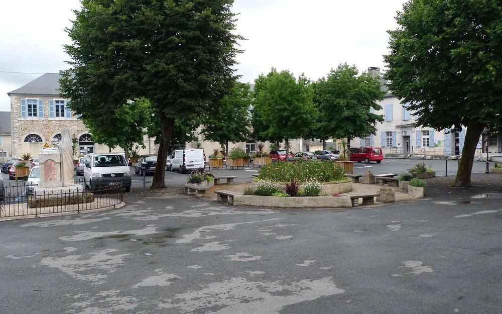 Rond-point d'une petite ville avec des arbres, des parterres de fleurs, des voitures garées et des bâtiments en pierre par une journée nuageuse