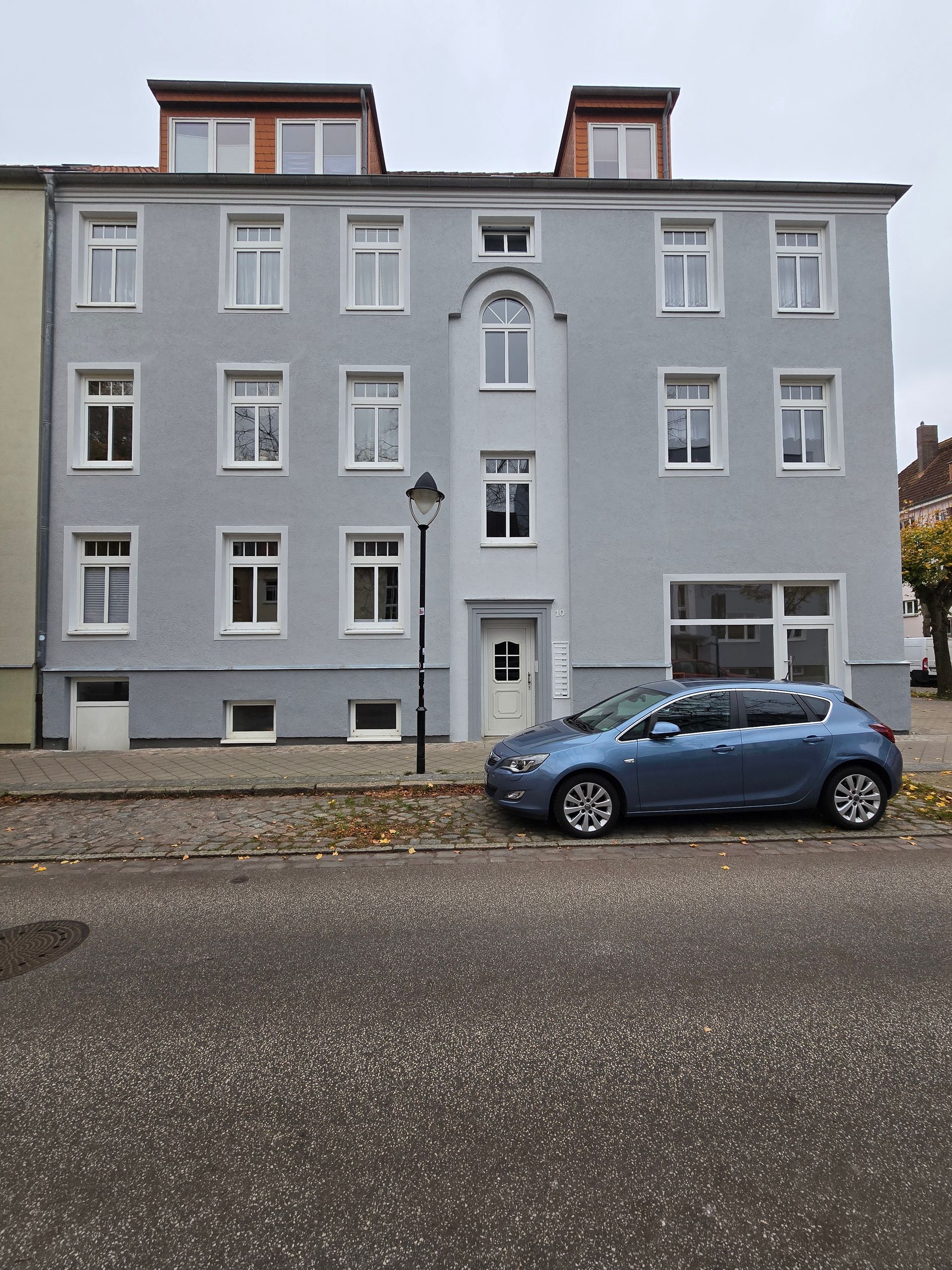 ein großes Gebäude mit vielen Fenstern in grau, im Vordergrund ein blaues Auto neben einer Laterne