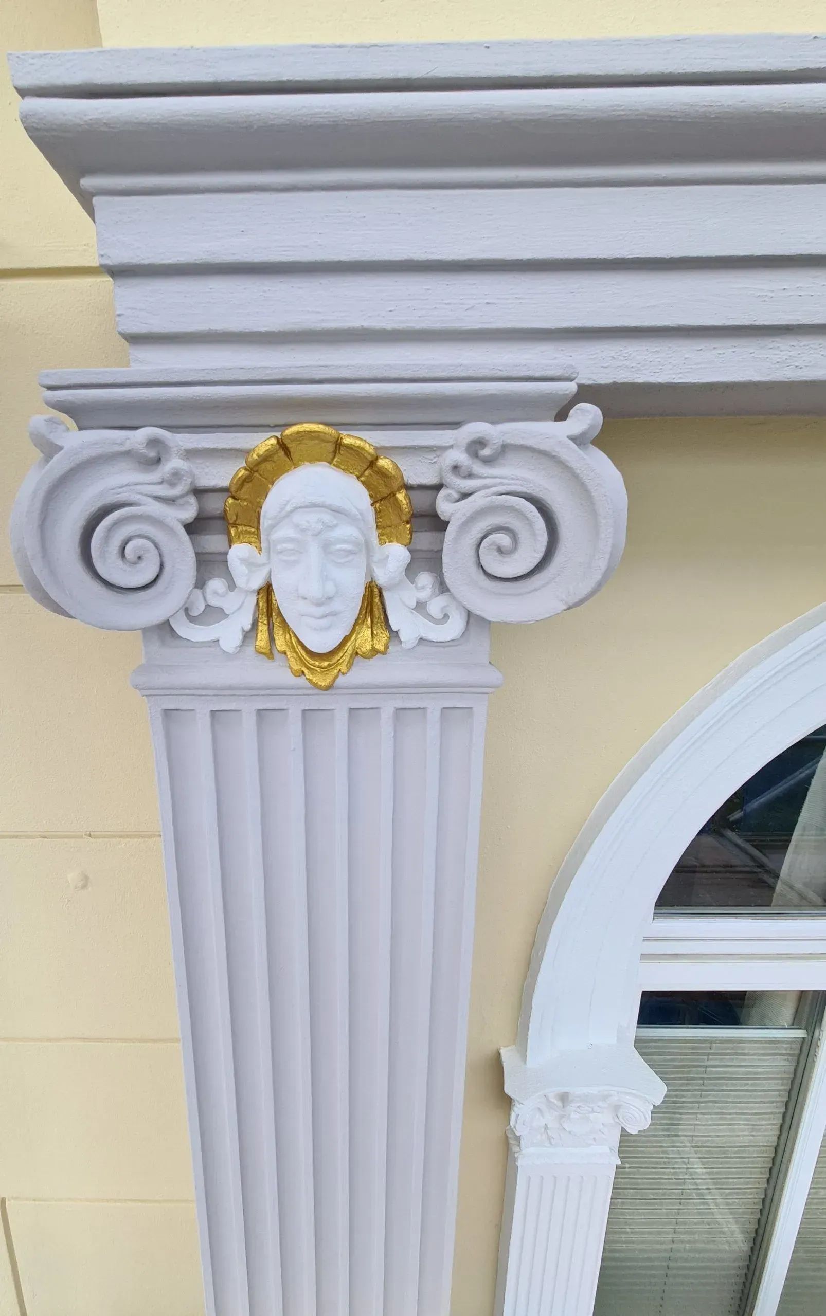 Ein detailreiches Fassadenelement im klassischen Stil. Eine weiße Säule mit einer Maske mit goldenen Akzenten. Als Maler bei Rostock reinigen uns sanieren wir Fassaden und Fassadenelemente.