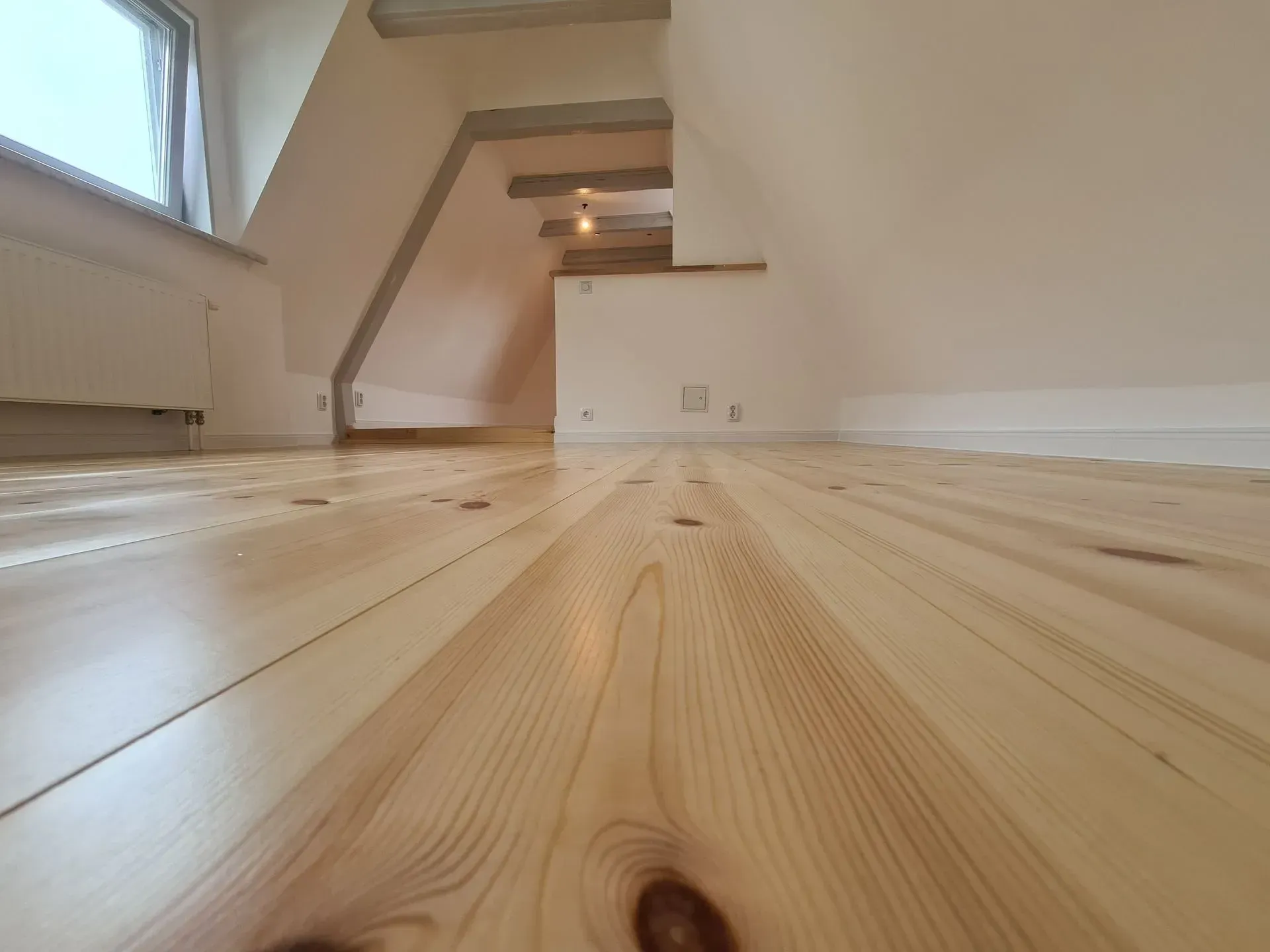 Ein frisch renovierter Dachgeschossraum mit hellem, naturbelassenem Dielenboden aus Holz. Die Wände sind in einem zarten Cremeton gestrichen. Als Maler bei Rostock verlegen wir Fußböden verschiedenster Art.